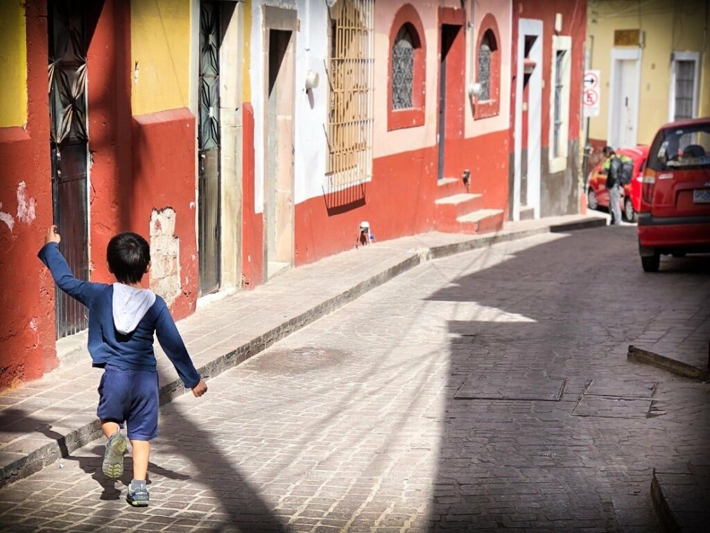 A boy walks through a colonial style street in Guanajuato, Mexico. The road and sidewalk are stone and brick, and the buildings are cement, with red, orange, and yellow paint. Cultural immersion is one of a many gap year ideas for families.
