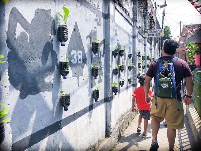 A man and a young boy walk through a small walkway in Yogyakarta, Indonesia, where travelers can learn Indonesian basic words. To the left of them, they pass a wall decorated with plastic bottles that have been converted into planters, and paintings of shadow puppets on the wall.