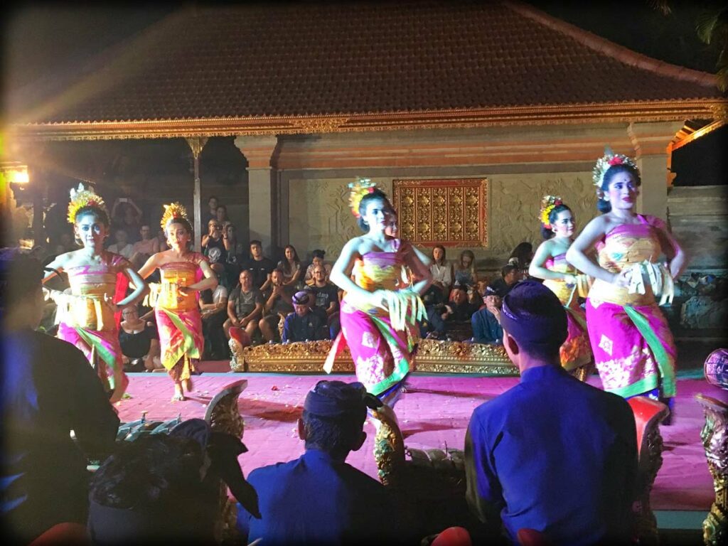 Dancers at the Ubud Royal Palace, one of the popular Ubud activities to do. Six dancers dance in the middle of a stage, in the foreground are gamelan players, facing the dancers and away from the camera. In the background are audience members watching the dancers.