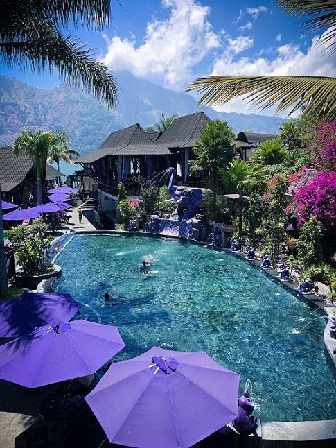 A Bali hot springs pool at Toya Devasya. Purple umbrellas line the left side of the pool. Purple elephant fountains line the right side of the pool, spouting water into the pool. In the background are palm trees and Mount Batur.