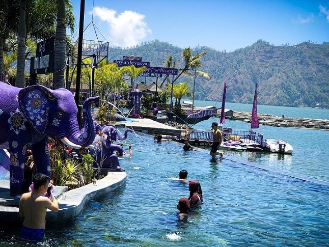 Swimmers in an infinity pool at Toya Devasya Bali hot springs. On the left are purple elephant fountain statues pouring water into the pool. In the background is Lake Batur, with hills in the far background.
