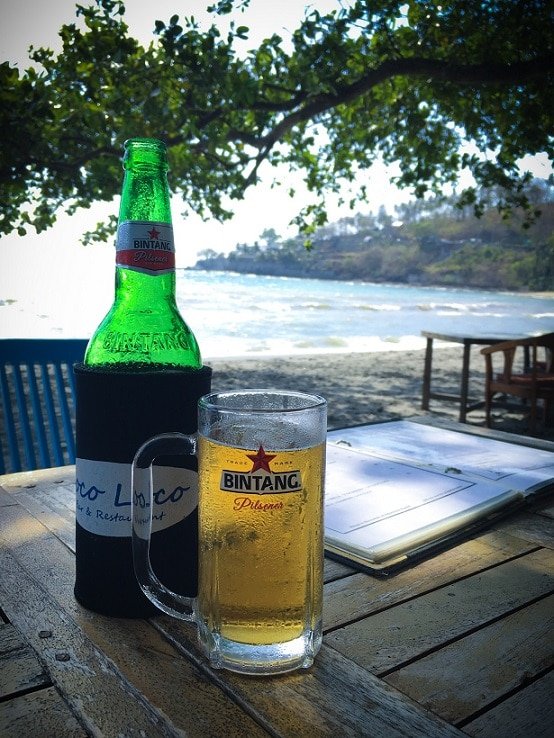 A bottle of Bintang beer with a glass of beer on a table at the beach, one of the popular things to do in Lombok.