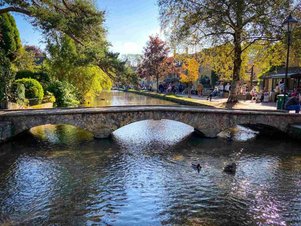 A footbridge over a river at Bourton-on-the-Water, England with ducks in the foreground, trees to the left, and a sidewalk lined with stores in the background