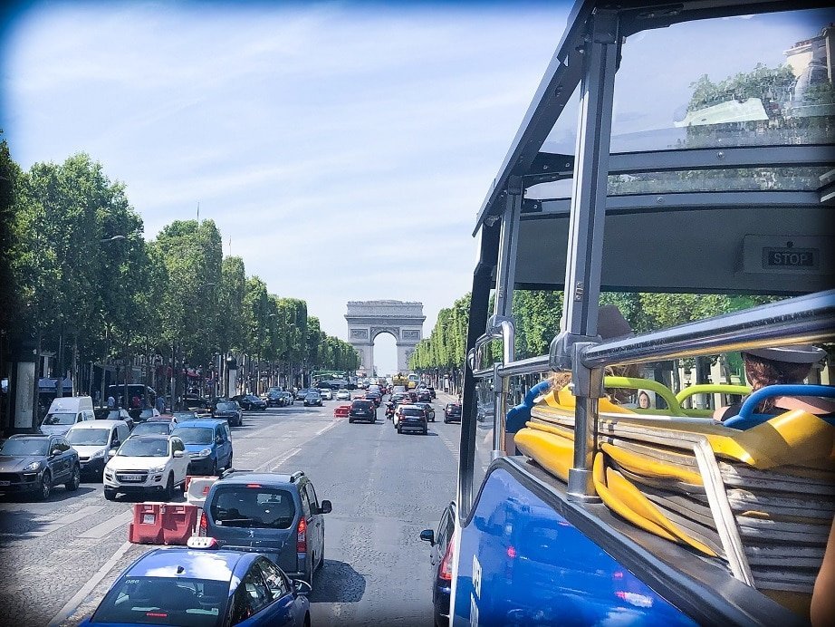 A view of Arc de Triomphe during a Paris 3 day itinerary. In the foreground corner is an open top tour bus. In the background is Arc de Triomphe, with the tree lined road filled with cars leading up to the arc.