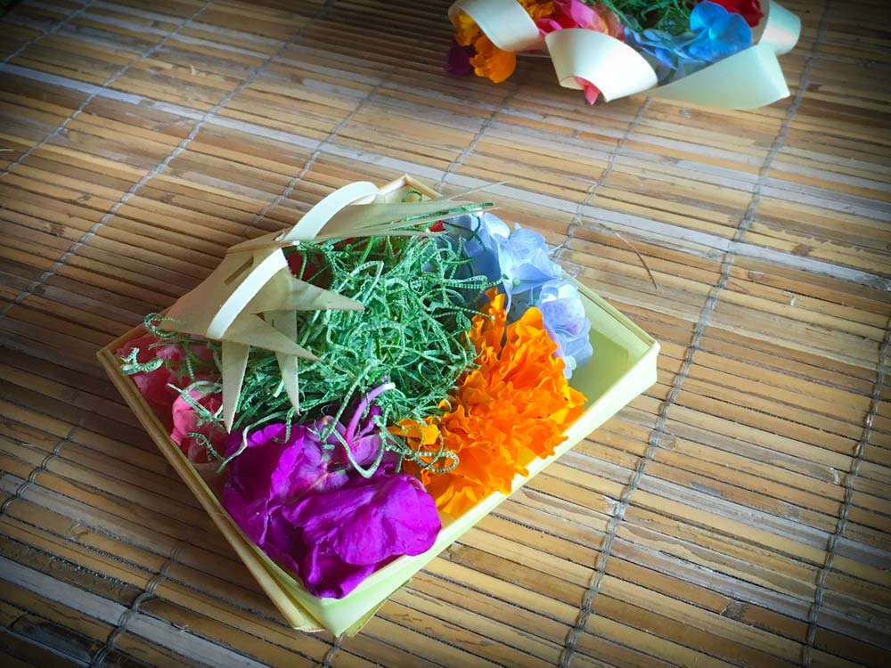 A traditional Balinese Hindu offering made as part of an excursion tour of Ubud activities. A small square container made with palm leaves, with pink, purple, orange, and blue flowers, along with green moss.