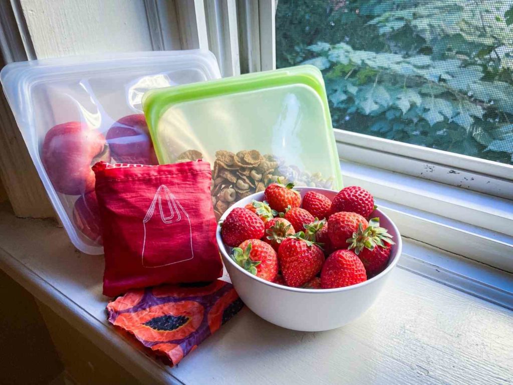 An assortment of eco friendly travel products next to a window: silicon bags, nylon tote bag, beeswax wrap, and a bowl of strawberries.