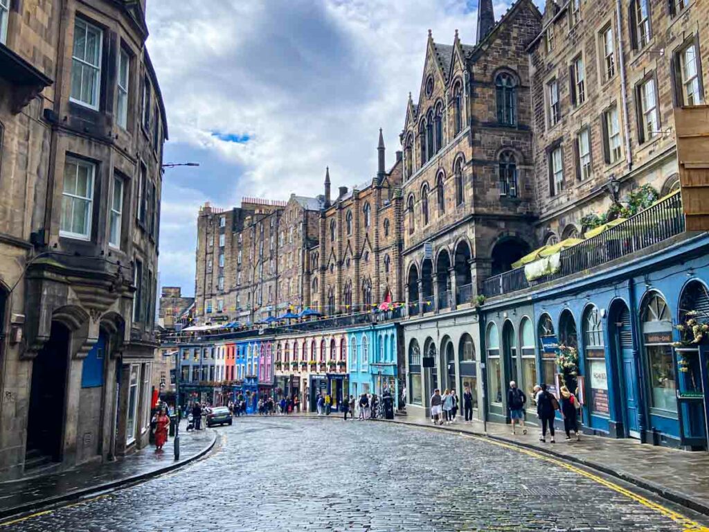 A view of Victoria Street in Edinburgh with buildings along a curve. The lower floor are bright colorful storefronts and the upper levels are a tan stone in old fashioned Victorian style architecture. People are walking on the sidewalk. This photo was taken during an Edinburgh 3 day itinerary.