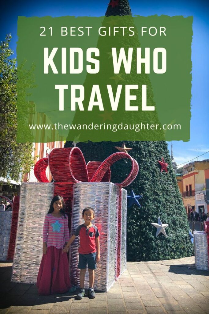 21 Best Gifts For Kids Who Travel | The Wandering Daughter | Pinterest image of two kids standing in front of a giant Christmas box and Christmas tree with text overlay
