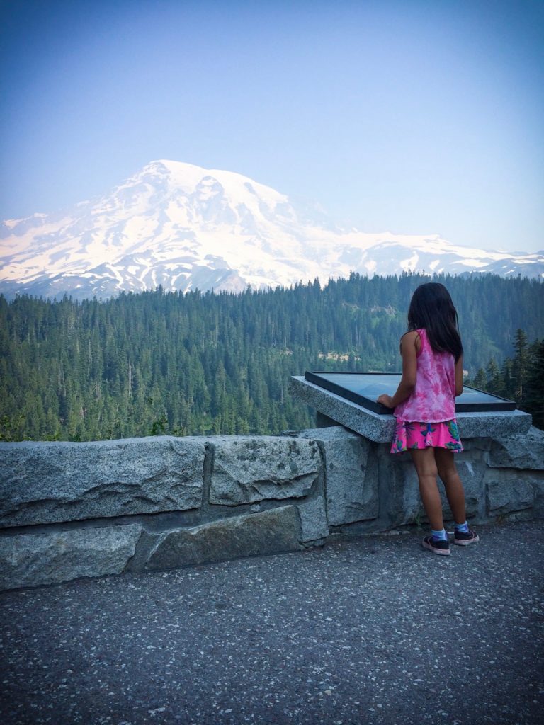 A young girl taking a break from Mount Rainier hikes and looking at Mount Rainier at an overlook at Mount Rainier National Park