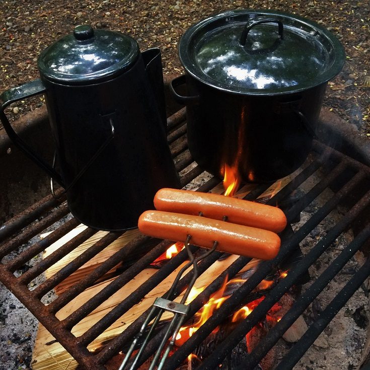 Hot dogs cooking on a campfire grill, with camping kettle and pot, during camping with a toddler