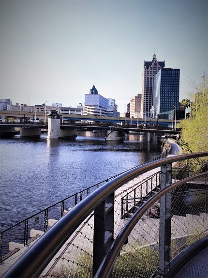 A river in Milwaukee, Wisconsin, with a sidewalk along the river. In the background are buildings in a city. The riverwalk is one of the popular Milwaukee activities for families.