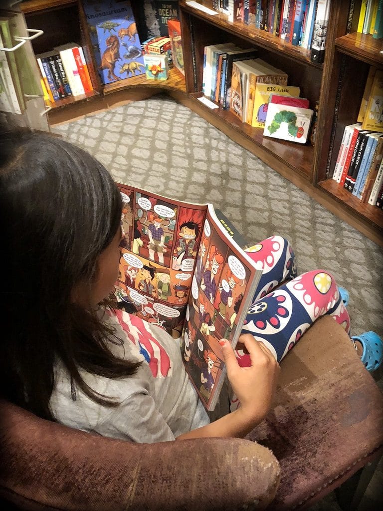 A young girl sitting in an arm chair in a book store, reading a graphic novel and using books as worldschooling resources.