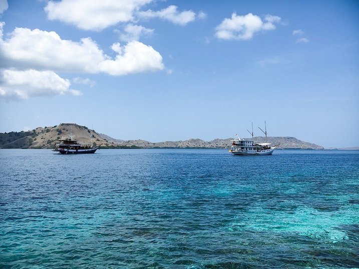 Two boats on the ocean during a Komodo tour. In the foreground is water with coral reefs, in the background behind the boats are mountains and sky.