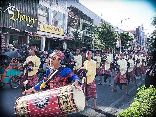 A musical group dressed in traditional Indonesian clothing perform on the street in Yogyakarta, where people can learn Indonesian. A drummer leads the procession, along with men in cymbals.
