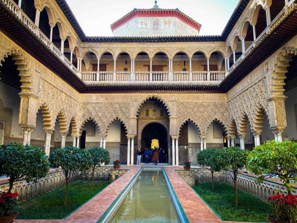 A courtyard with a pool of water in the middle surrounded by small trees and Arabic style arches and columns
