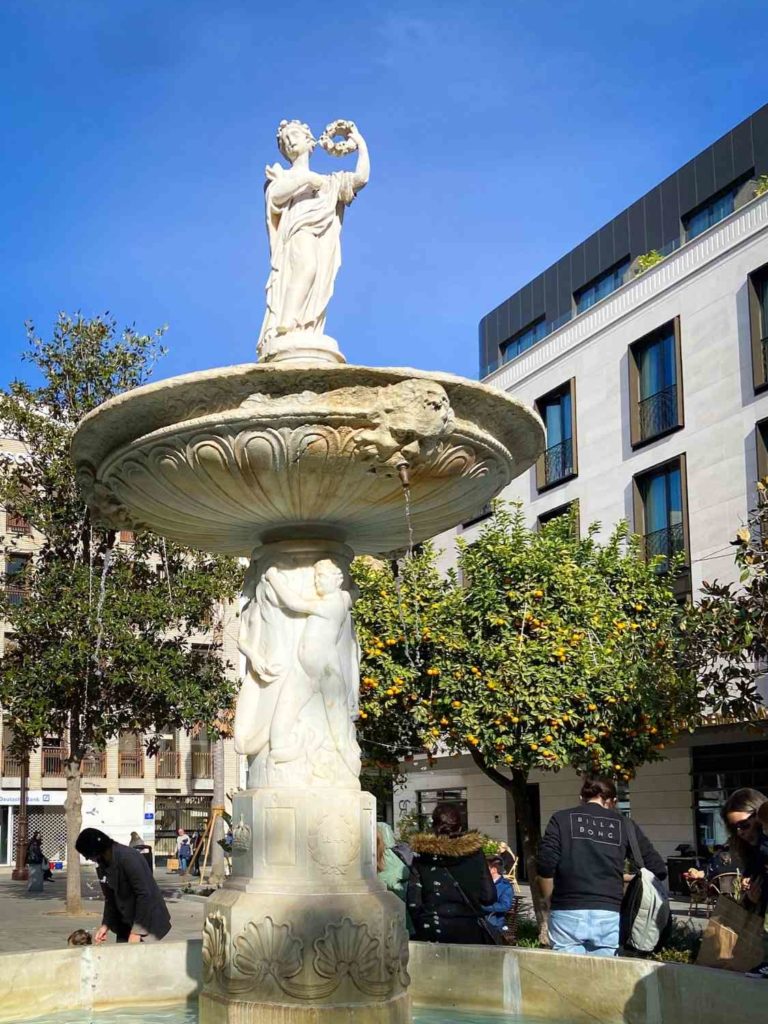 A circular fountain with a figure of a woman at the top and buildings in the background