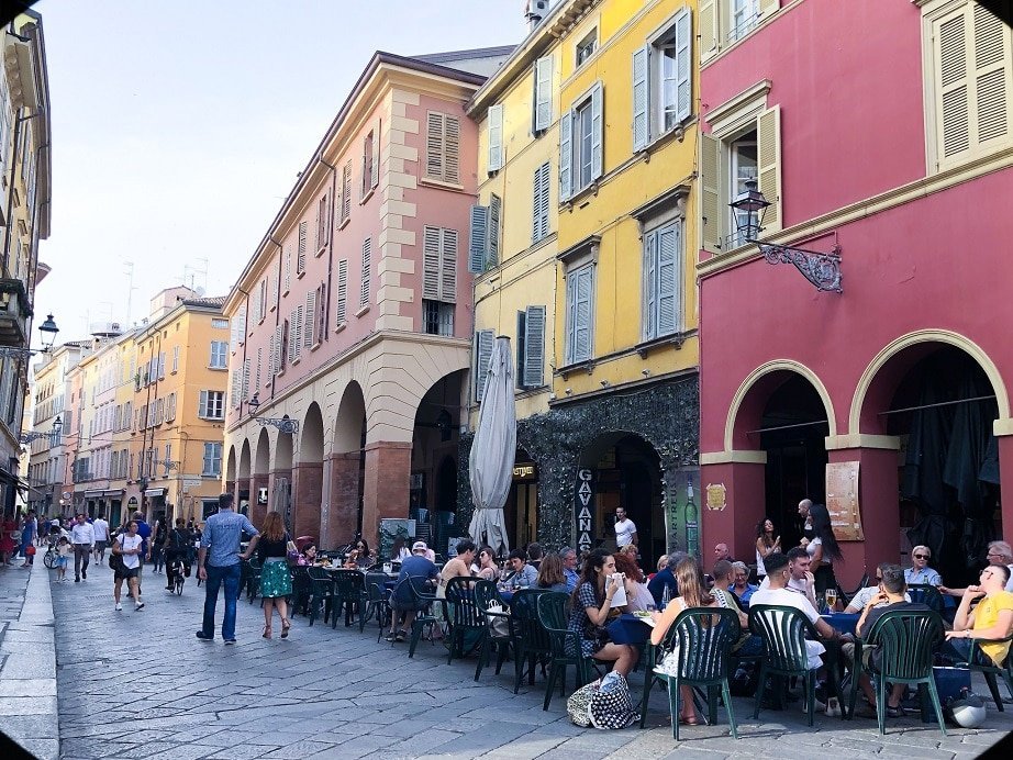 A street in the Centro Storico of Parma Italy. Brightly colored buildings in 17th century style architecture, with cafes on the sidewalk in front of the buildings. An example of what to do in Parma.