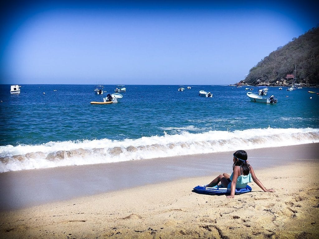 A young girl sitting on the sand on the beach, her back to the camera and looking out onto the water. In the the distance are fishing boats scattered throughout the water.