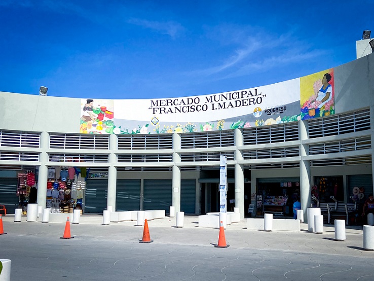 The entrance to the Municipal Market, one of the things to do in Progreso. A curved building facade, with a sign on top saying, "Mercado Municipal Francisco I.Madero". Small shops line the front of the building. Orange cones are in a line the front of the entrance to the building.