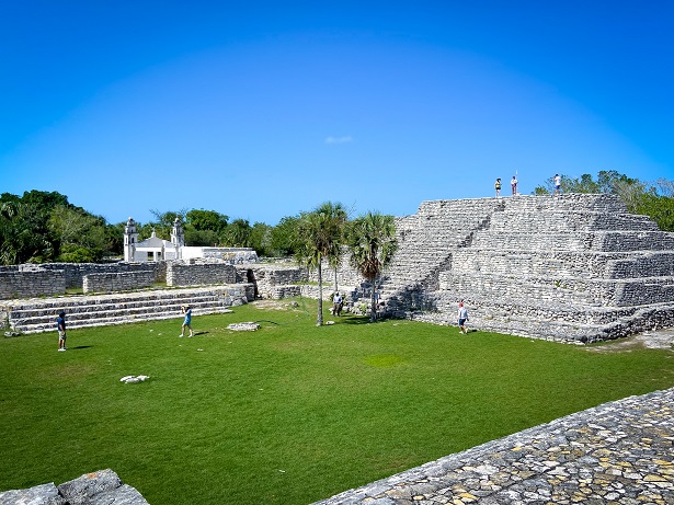A stone pyramid with palm trees in front of it and next to a platform made of stone. A grassy field sits in front of the pyramid and stone platform. The pyramid is located in archaeological site, Xcambo, one of the things to do in Progreso, Mexico.