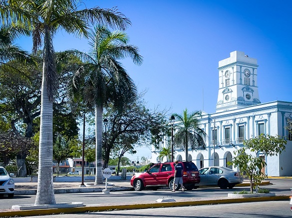 A blue and white colonial building in Progreso, Mexico with cars parked in front of it and palm trees next to it.