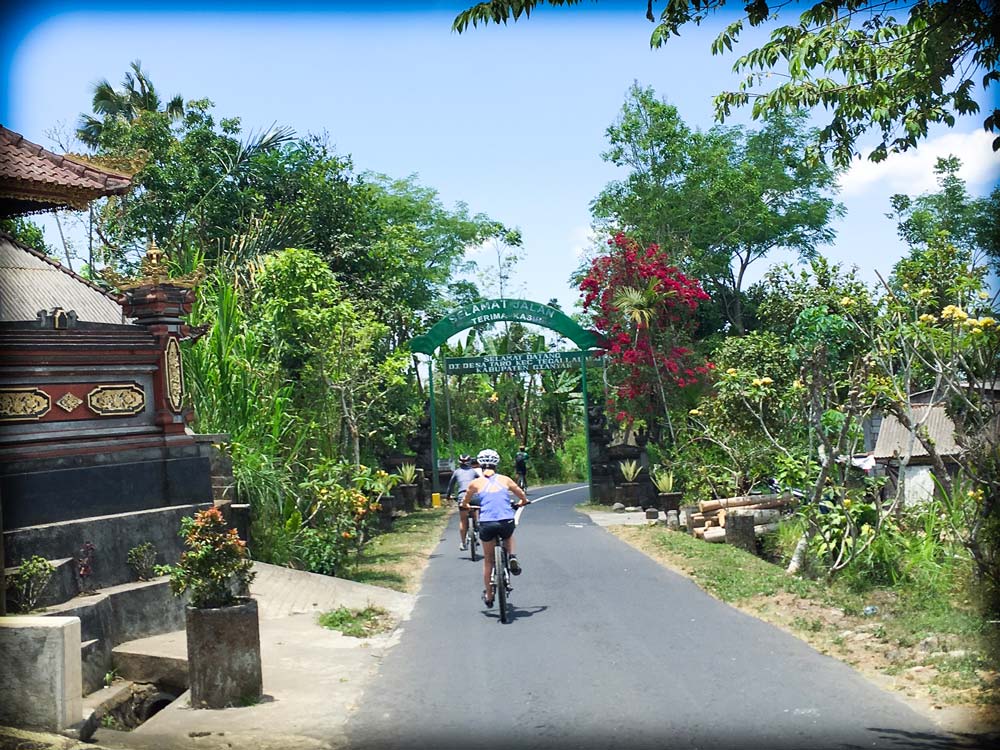 A bicyclist rides along a paved road in Bali, as part of downhill cycling Ubud activities. The bicyclist is biking away from the camera, so we see their back. To the right of the bicyclist are trees and plants, and to the left of the bicyclist is a traditional Balinese building in the foreground, and trees in the background. In front of the bicyclist is an arch across the road with words written on it.
