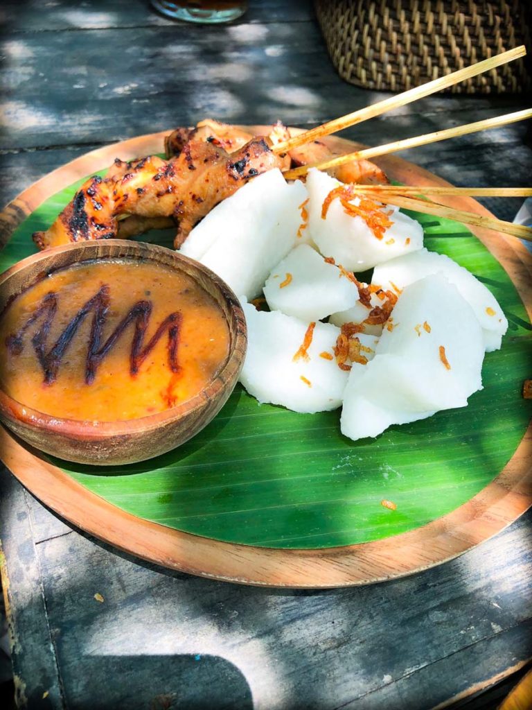 Satay ayam with nasi tipat, a popular meal after doing Ubud tourists activities. A bamboo plate with a banana leaf liners. On top is a stack of rice pressed into cubes, with skewers of chicken satay. On the side of the plate is a small wooden bowl with peanut sauce.