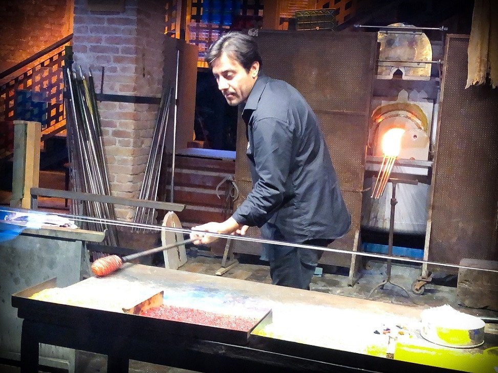 A glassblower demonstrates how to make blown glass during a personal Venice walking tour. He rolls hot glass, attached to a long hollow metal tube, on top of bits of crushed glass on a table.  