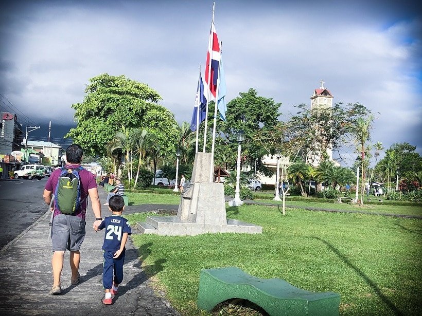 A man holding a young boy's hand, showcased on family blogs, as they walk through a small town in Costa Rica. They are passing a green field with a flag pole in the immediate distance, and a tower behind trees in the distance.