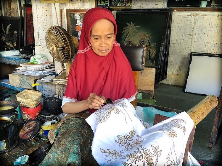 An older Indonesian woman in traditional headscarf prints a batik art pattern with wax on fabric, using a batik writing tool, at a batik making workshop in Yogyakarta, Indonesia.