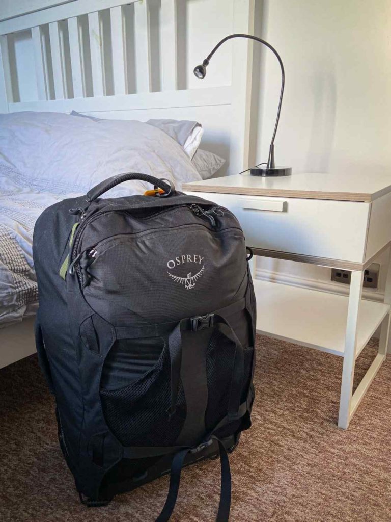 A black Osprey Farpoint 36, an option for the best backpack for travel with kids, standing upright in front of a bed and next to a white side table with a desk lamp.