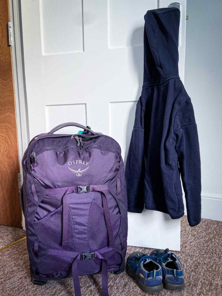A purple Osprey Fairview 36, an option for best backpack for travel with kids, in front of a white door with a dark blue sweatshirt hanging on the handle and a pair of blue Keen sandals next to it.