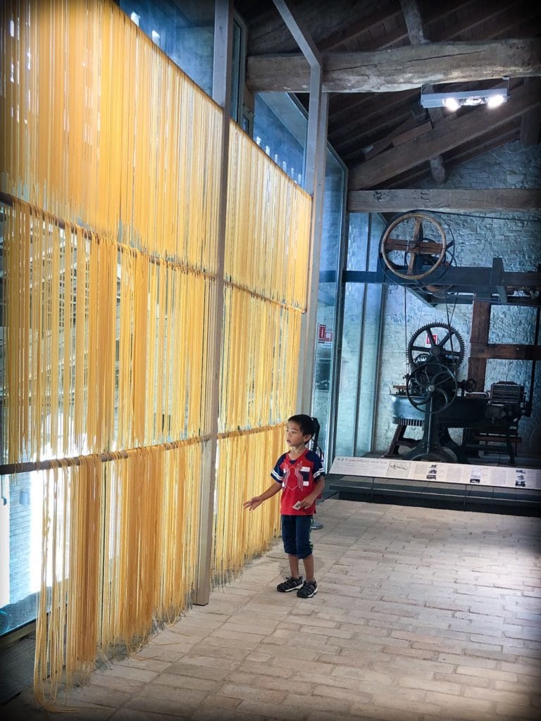 A child what is worldschooling in a pasta museum in Italy looking at a giant drying rack with noodles drying on them. An industrial pasta machine is on display behind him. 