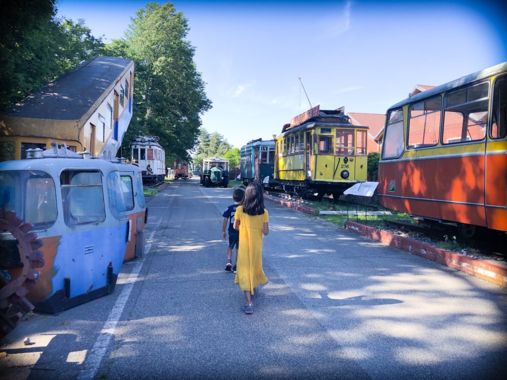 A boy in blue clothes and a girl in a yellow dress walk along a paved road at Volandia museum in Milan, Italy. The road is lined with old trams, trolleys, and train.