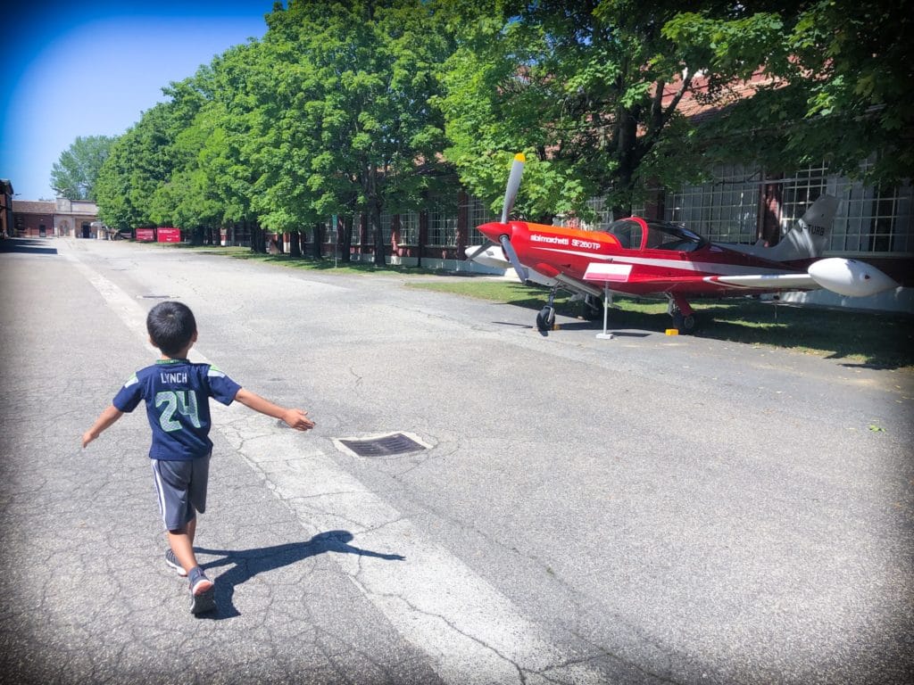 Young boy walking through the outdoor grounds of Volandia museum of aviation in Milan, Italy, passing by a small red propeller plane.
